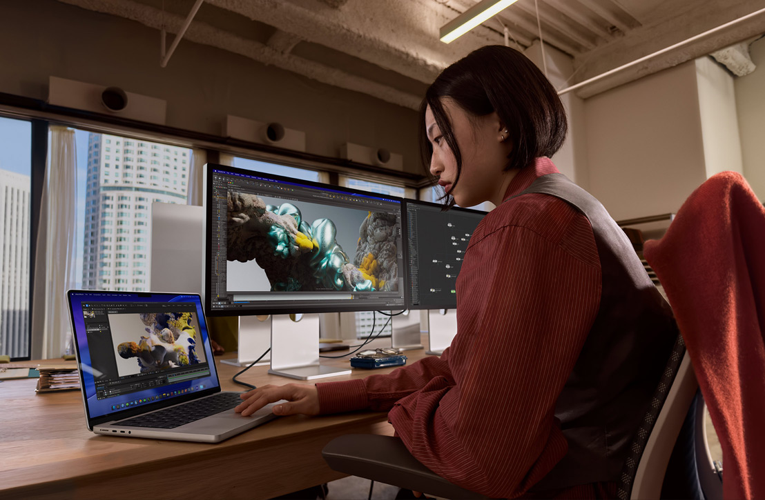 Una persona seduta a una scrivania che lavora con un MacBook Pro collegato a un monitor esterno; entrambi gli schermi mostrano un rendering 3D, dalla finestra dietro al monitor si notano alcuni edifici