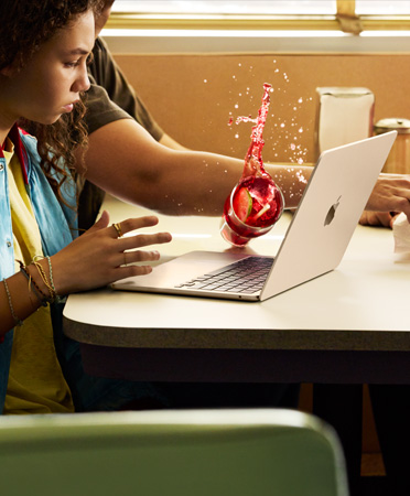 Una persona en un restaurante da vuelta una bebida con hielo sobre un MacBook abierto al estirar el brazo para tomar algo de la mesa