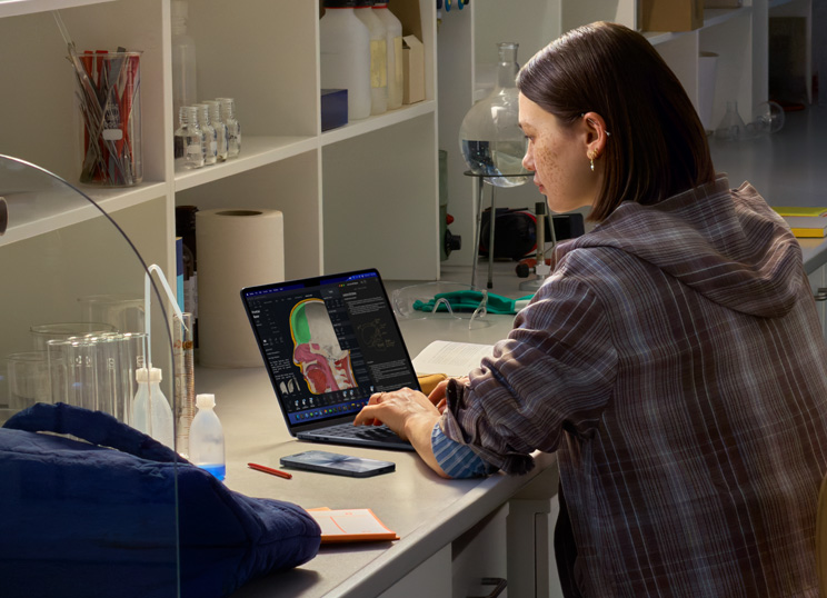 Uma pessoa sentada em um laboratório usando um MacBook, com a tela exibindo vários apps