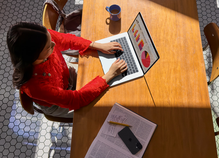 Uma pessoa usando um MacBook em uma mesa, com uma caneca, um smartphone e papéis por perto