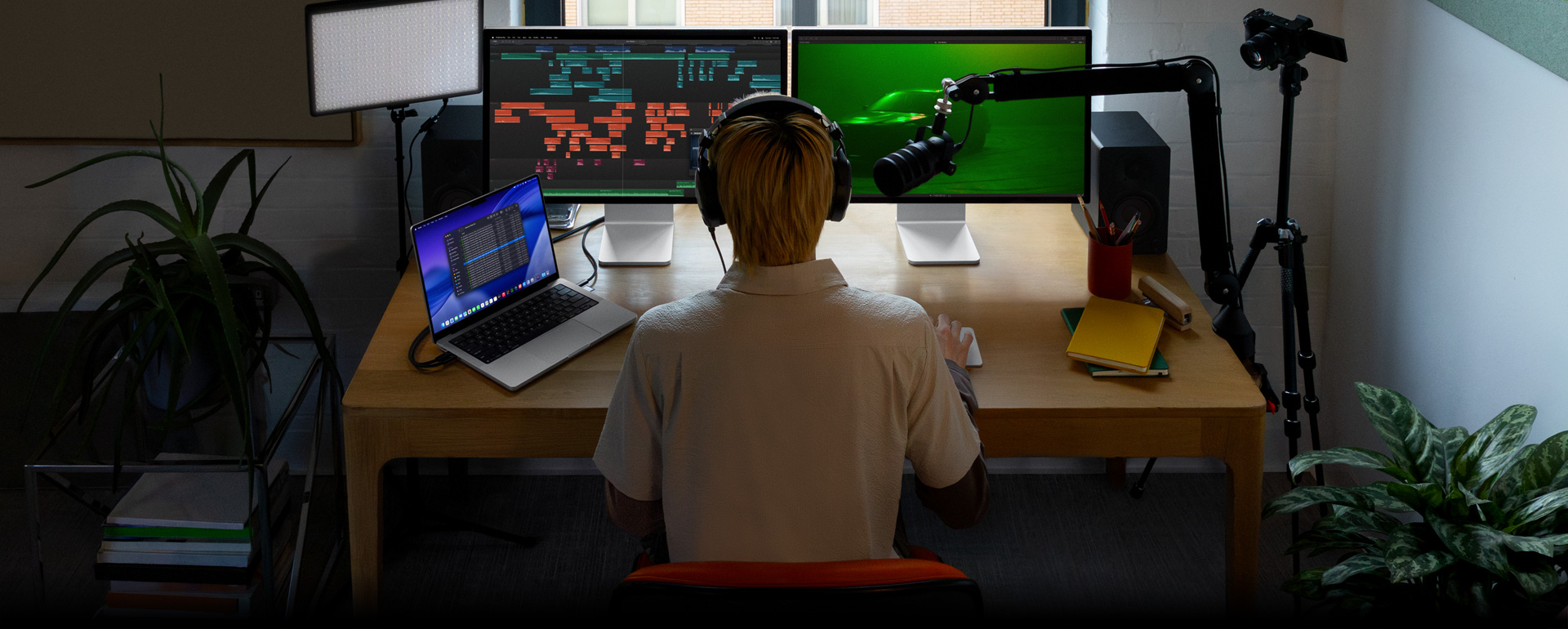 Persona sentada en un escritorio con un micrófono y audífonos frente a tres dispositivos: un monitor Studio Display para la Mac que muestra herramientas de edición de audio y clips de sonido en distintos bloques de color, un segundo monitor Studio Display para la Mac que muestra un auto en una escena con iluminación ambiental verde, y una MacBook Pro de 16 pulgadas con una larga lista de tomas en un proyecto de Final Cut Pro