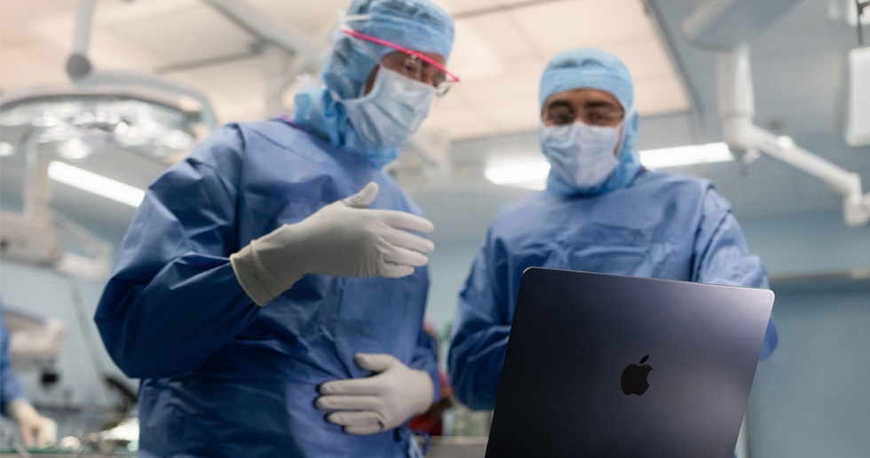 Dois cirurgiões na sala de operações a olhar para um MacBook, um faz gestos com a mão enquanto o outro observa