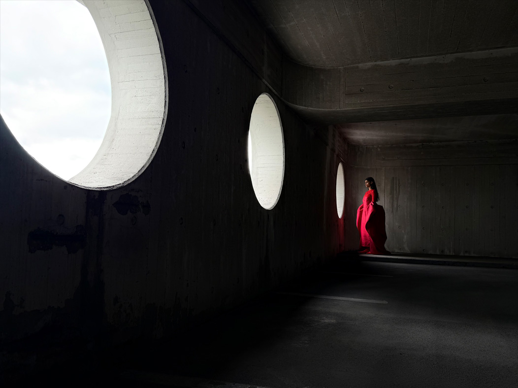 Una mujer con un largo vestido rojo de pie en medio de una habitación vacía mientras la fotografían. La distancia focal de la imagen se va alternando en la app Fotos para mostrar las capacidades del sistema de cámaras Pro: 28 mm