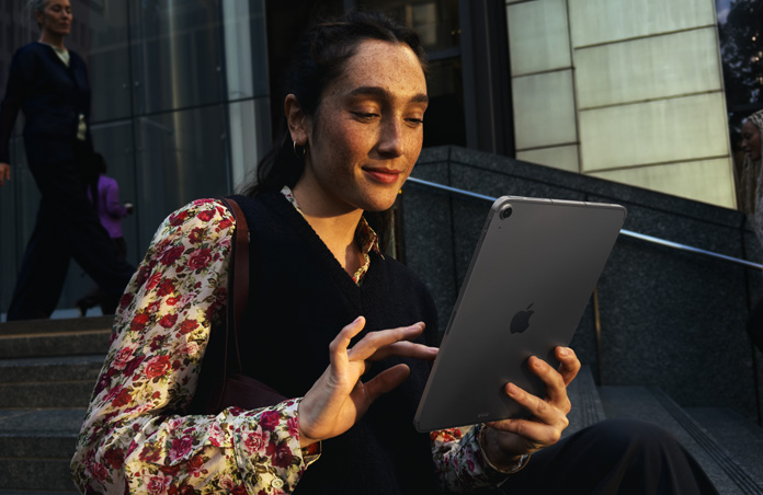 Una persona sentada en las escaleras afuera de un gran edificio usa un iPad Air de 11 pulgadas gris espacial, con una conexión Wi-Fi que se mantiene estable mientras alterna entre diferentes redes